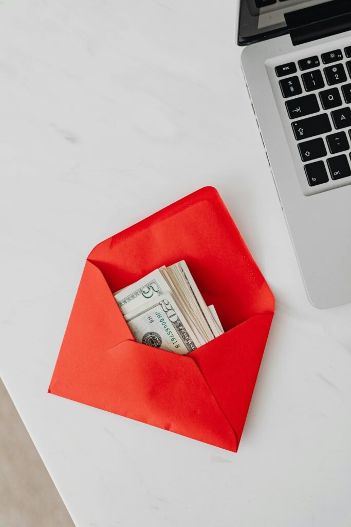 Top view of dollar bills in a red envelope next to a laptop on a white desk.